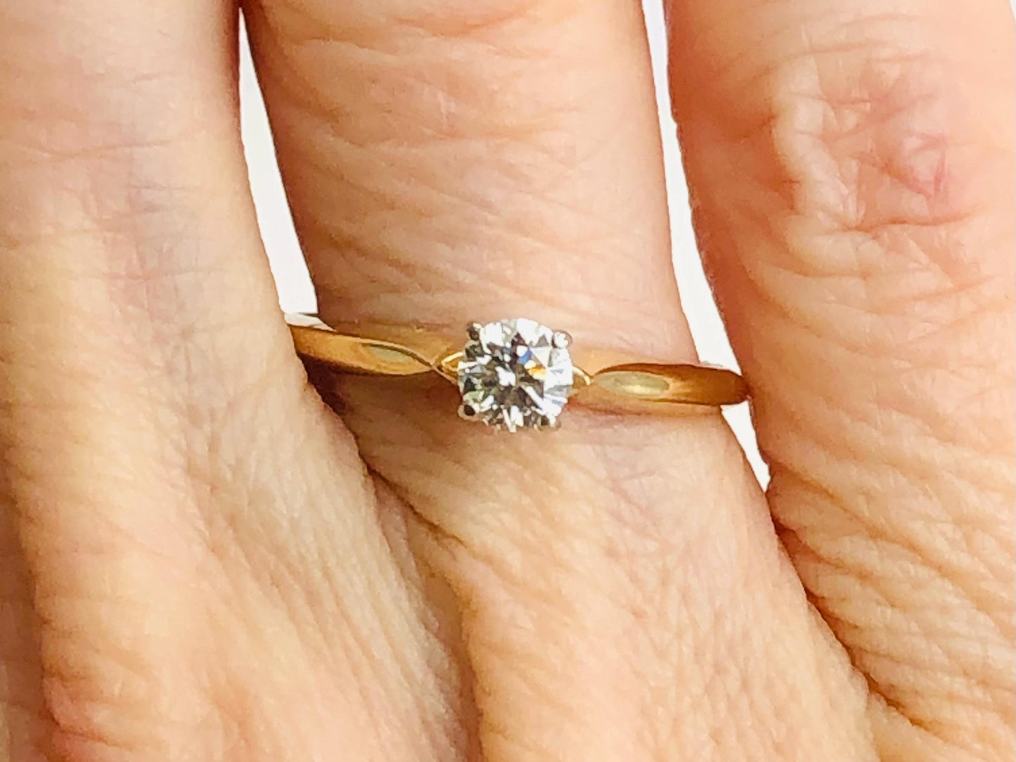May include: A solitaire diamond engagement ring with a round-cut diamond. The diamond is set in a four-prong setting on a delicate yellow gold band. The ring is simple and elegant, perfect for a classic engagement ring.